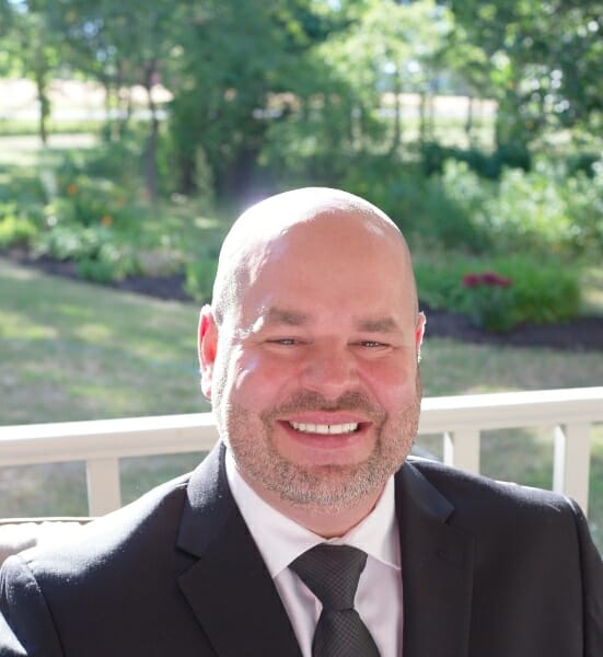 derek-lunau-western-pride-woodworks - Western Pride Woodworks Smiling bald man in a dark suit and tie on a sunny outdoor deck.